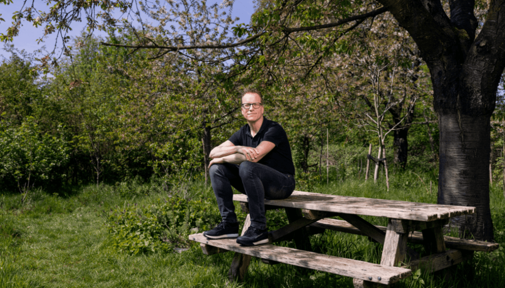 Een man zit op een picknicktafel met zijn voeten op het bankje. Hij heeft zijn armen over elkaar en kijkt neutraal in de camera. Om hem heen staan fruitbomen en op de voorgrond is gras.