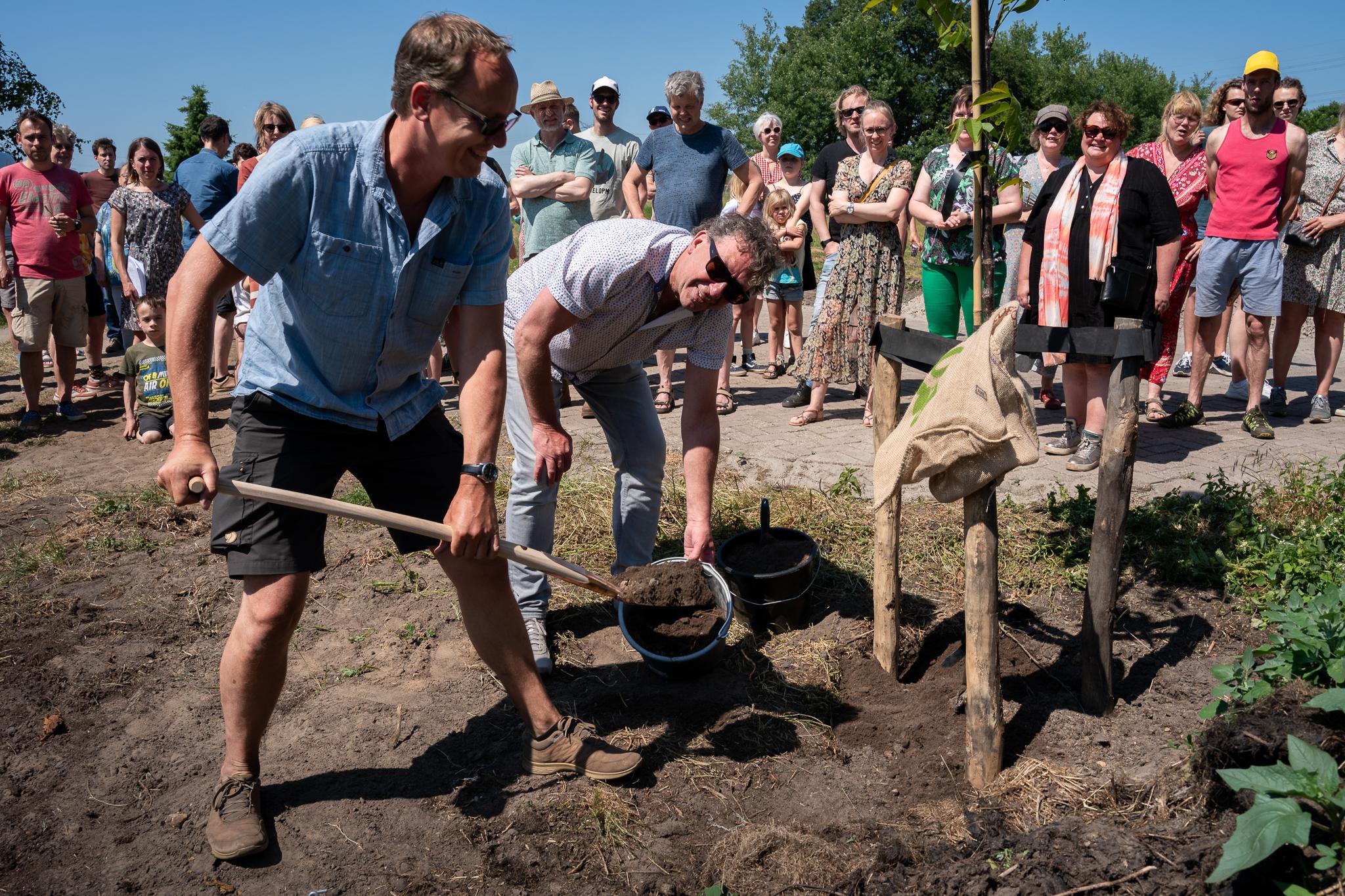 Een rij mensen in zomerse kleding met op de voorgrond twee mannen die een kuil dichten waarin ze zojuist een boom hebben geplant.
