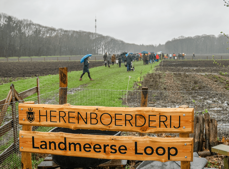 Een aantal akkers in de regen en daarop een groep mensen met paraplu's. Op de voorgrond een houten bord met de woorden Herenboerderij en Landmeerse Loop.